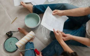 Dwie osoby siedzą na podłodze z wydrukiem planu, obok puszka farby, wałek i miarka